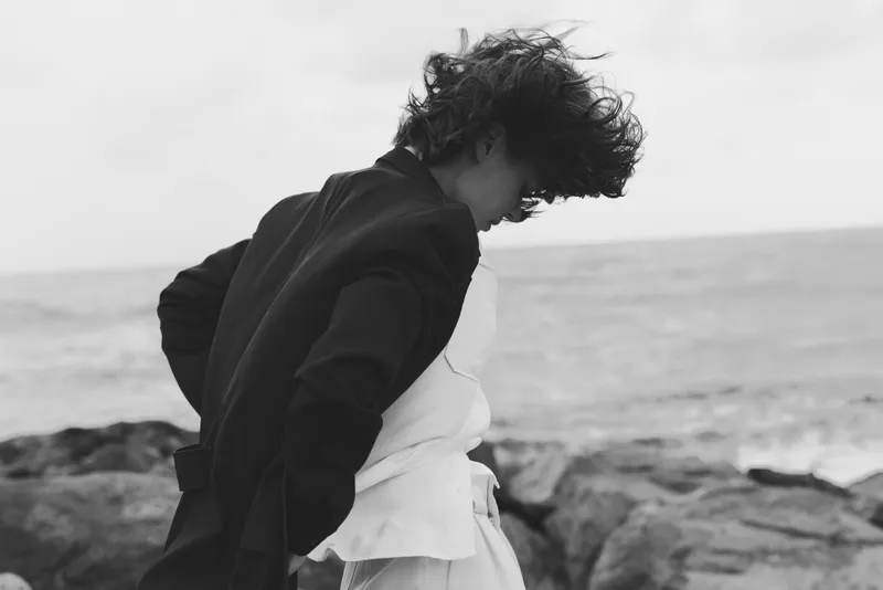 Black and white embrace along coast, couple moment by the sea, wind in hair, flowing garments, moody documentary feel, outdoor atmosphere, intimate connection.