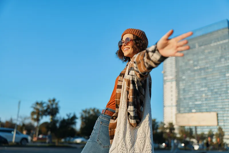 woman smile city carefree fashion sunlight, urban portrait of a joyful young woman with arms out, layered outfit and sunglasses, authenticity, candid lifestyle, golden hour glow.