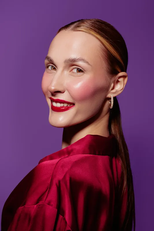 Smiling woman with sleek hair and red lipstick wears shiny red satin blouse against purple background. Portrait with elegant makeup and confident expression. Concept of beauty and fashion.