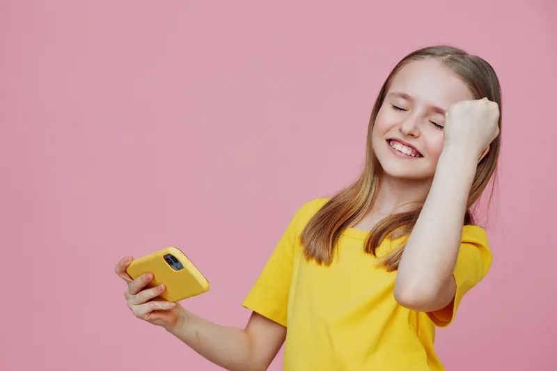 Happy girl celebrating success with a smartphone, wearing a yellow t shirt against a pink background, showcasing joy and excitement in a vibrant and playful atmosphere