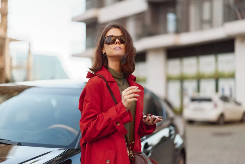 Confident young woman in a red coat stands next to a black car holding a smartphone and wearing sunglasses on a city street.