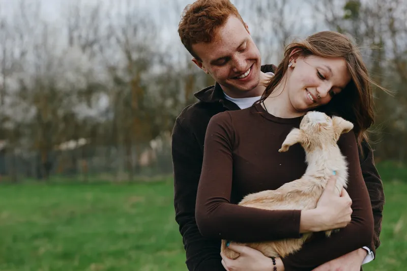 Cheerful couple outdoors, embracing with a small animal in a lush park, warm natural light, relaxed posture, joyful connection, soft breeze, vibrant greenery, candid lifestyle moment, sunlit and