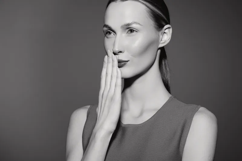 Portrait of a fair skinned Caucasian woman with sleek hair covering mouth with hand, looking thoughtful on grey background in studio lighting.