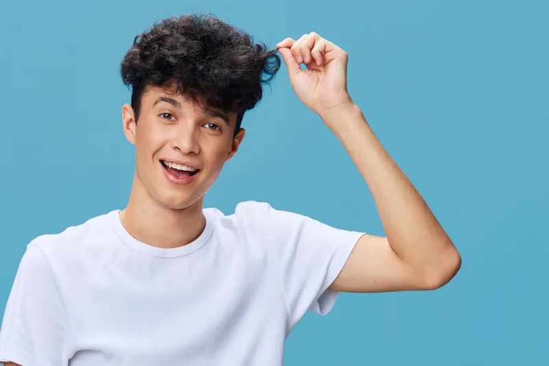smiling man with curly hair wearing white t-shirt standing against blue background expressing happiness and joy in casual style