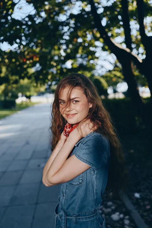 smiling woman in denim dress outdoors with long hair and natural lighting on tree-lined urban path during summer afternoon