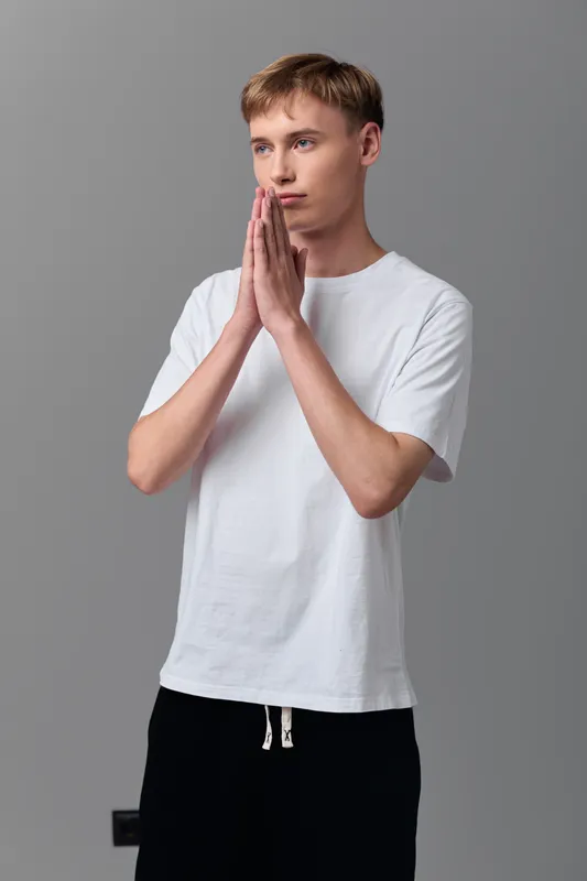 Praying gesture, hands pressed together, young man, white shirt, calm expression, meditation as he stands in studio, conveying focus, spirituality and inner peace in a serene moment