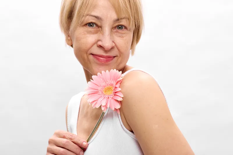 Woman smiling in studio portrait, holding pink flower near shoulder, soft light, gentle expression, closeup composition, clean white background, calm mood