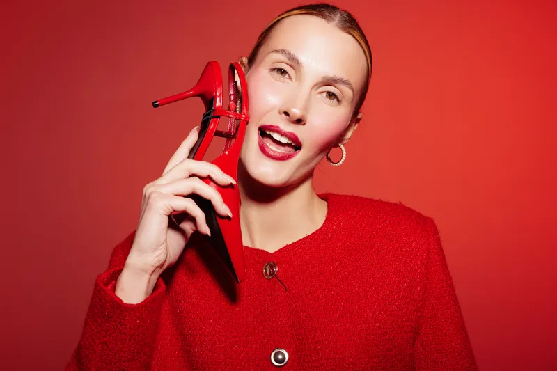 Smiling woman in red coat holds red high heel shoe near face against red background. Fashion concept with bright colors and stylish accessories in studio lighting.