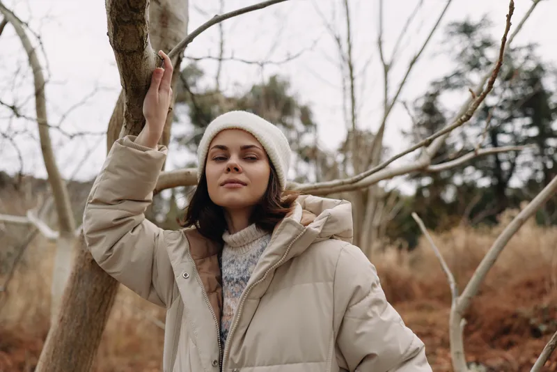 Winter portrait of a woman in a beige parka and knit hat, standing among bare trees in a cold outdoor setting with a confident expression.