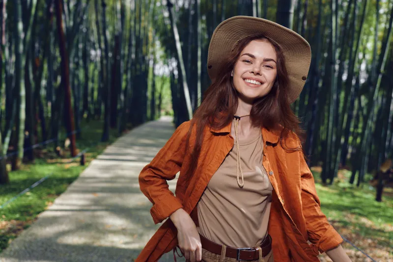 Woman smiling in hat and orange coat standing on path in a bamboo forest, portrait of a happy traveler enjoying nature and sunlight with trees and greenery around, casual carefree mood.
