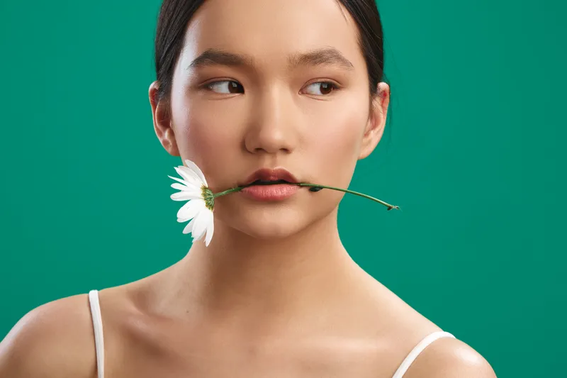 asian woman holds a flower between lips in a studio portrait, serene expression and clean aesthetic, soft lighting, minimal backdrop, highlighting beauty, balance, and subtle playfulness.