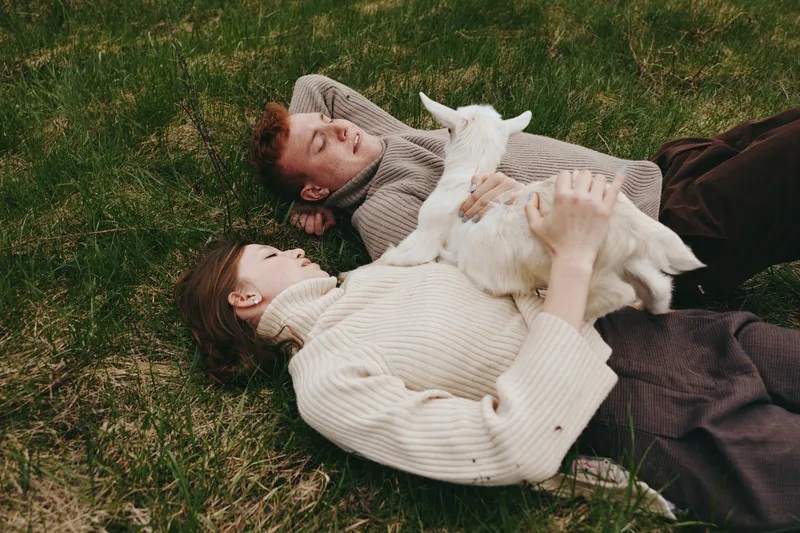 Young couple lying on grass with a white baby goat enjoying a relaxed moment in casual sweaters during a cozy outdoor lifestyle scene.