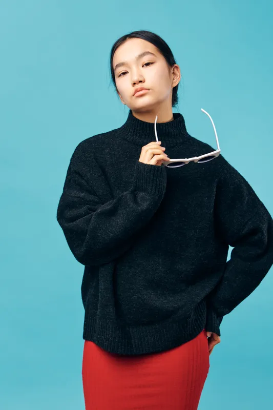 Young woman wearing black turtleneck sweater and red skirt holding white glasses with confident facial expression on blue background in studio portrait.