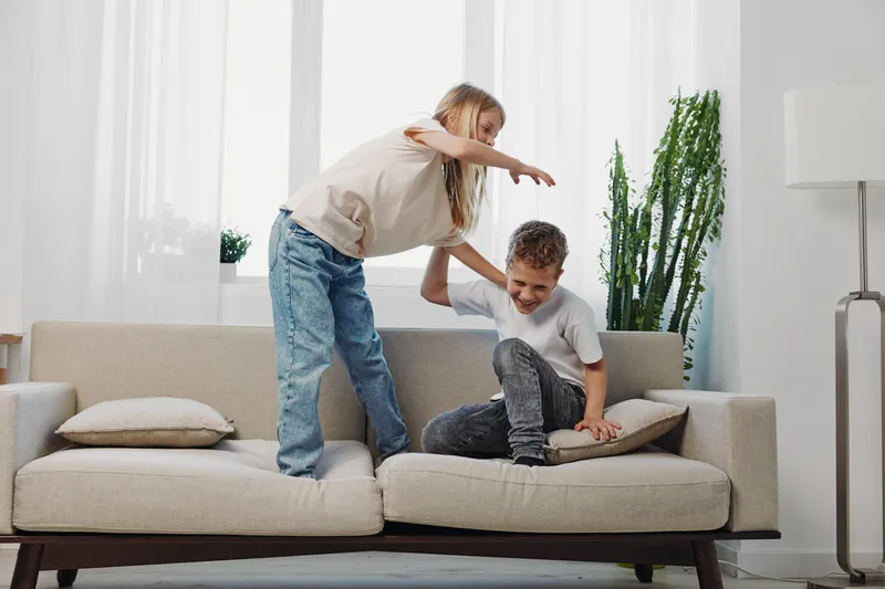 Two children playing and laughing on a beige sofa in a bright living room. The boy sits holding a cushion while the girl leans forward with arms outstretched. Home fun and sibling joy.