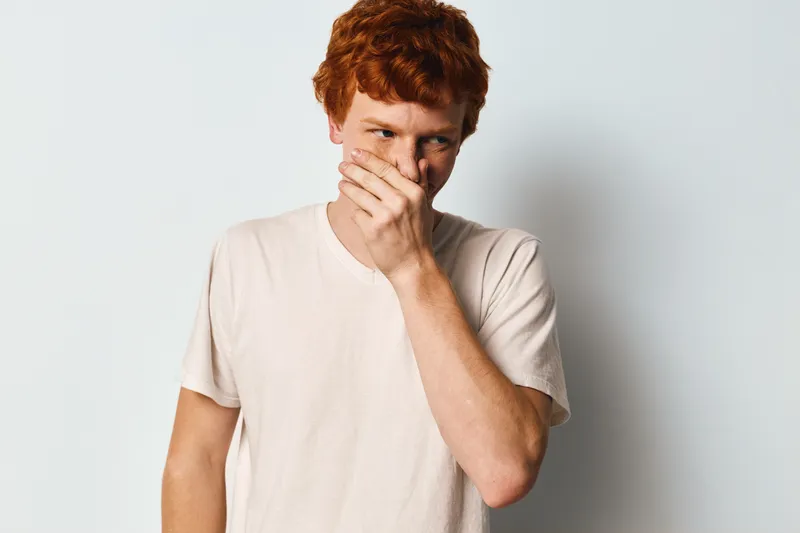 Young man with red hair and freckles wearing white t shirt covering mouth with hand in thoughtful or shy pose on light background with casual style and expression.