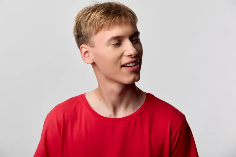 Young man in a red shirt turns to the side with a smile, casual studio lighting and a relaxed pose, clean background, everyday fashion and approachable mood.