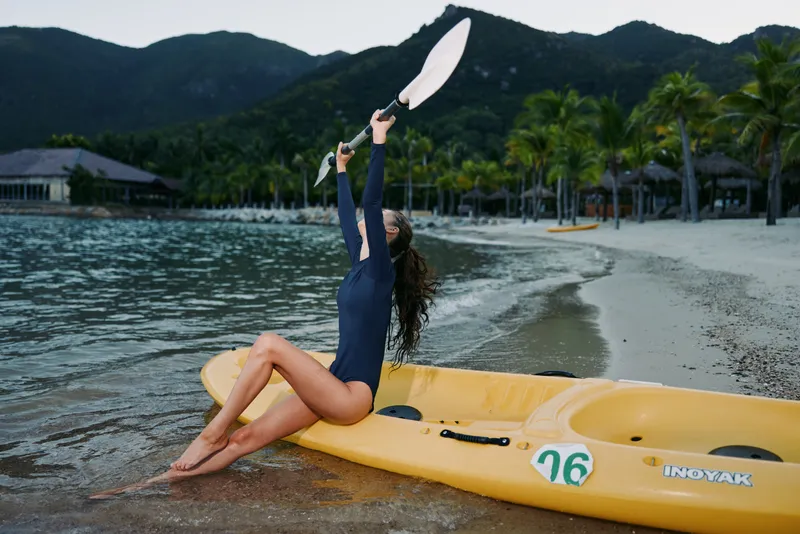 A female swimmer in a dark wetsuit sits on a yellow kayak at the shoreline, raising a paddle into the air as calm water and distant mountains frame the scene.