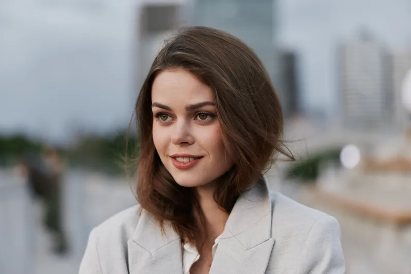 Confident businesswoman in a cityscape with a soft smile, dressed in a light gray coat, radiating professionalism and approachability, perfect for corporate branding.