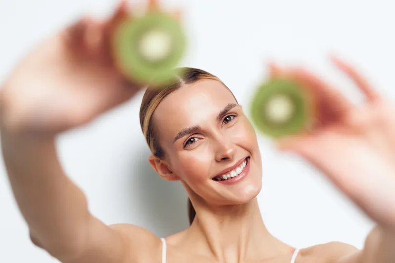 Smiling woman holding two halves of fresh kiwi fruit close to the camera. Bright studio lighting highlights healthy skin and joyful expression on a white background.