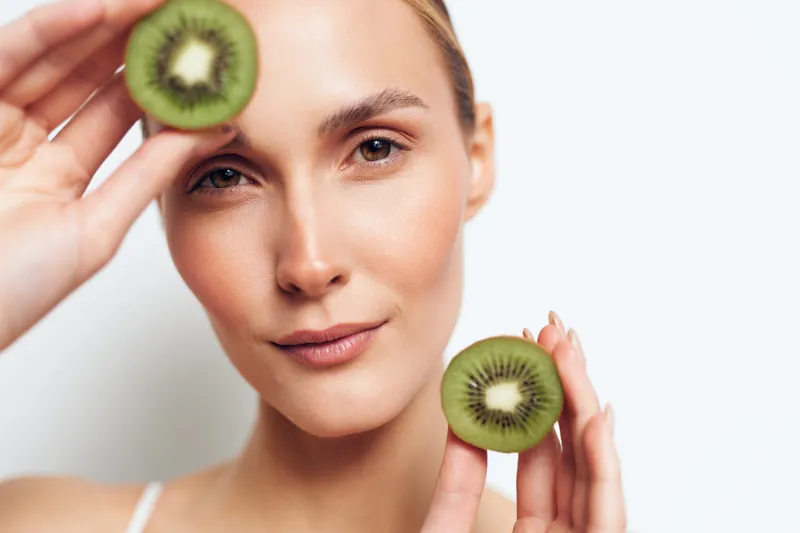 A young woman holds two kiwi slices near her face, presenting fresh fruit and a radiant complexion in a bright studio, creating a mood of healthy eating and wellness.