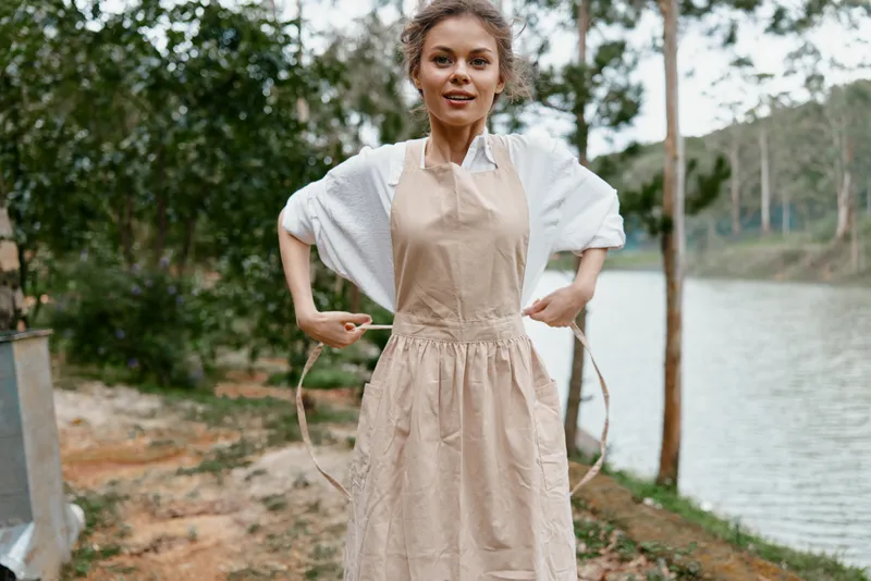 Young woman in an apron smiling by a serene lake, she embodies happiness, nature, and outdoor lifestyle with a light, earthy color palette