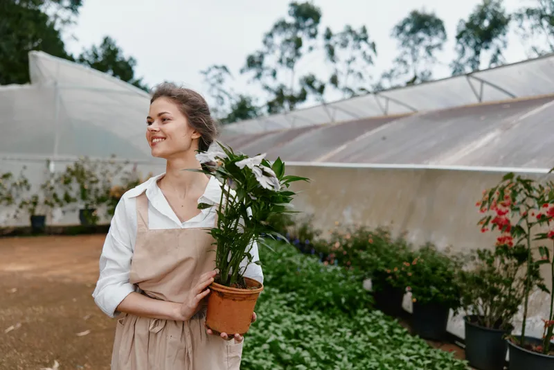 Young woman smiling while holding a potted plant in a greenhouse, showcasing gardening enthusiasm and vibrant greenery, ideal for seasonal themes and lifestyle content