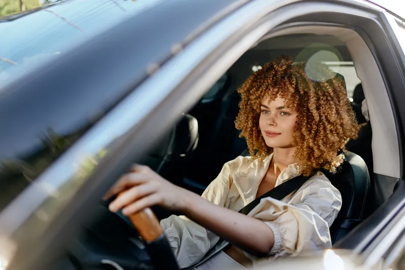 Young woman driving car with curly hair, wearing beige jacket, focused and calm, daylight, modern vehicle interior, natural light, casual lifestyle scene.