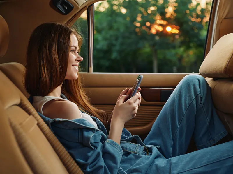 Young woman enjoying her phone in a cozy car during sunset, wearing casual denim outfit, exuding happiness and relaxation in a warm atmosphere.