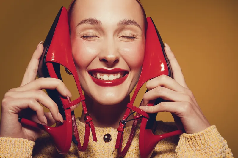 Happy woman with closed eyes holds red high heel shoes near face smiling. Bright makeup and cozy sweater create warm mood on yellow background. Concept of fashion and joy.