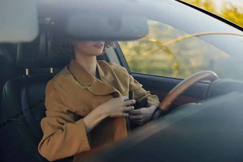 A woman sits in a car, adjusting her brown jacket, sunlight softly filtering through the window and casting a warm glow on her focused expression and the steering wheel