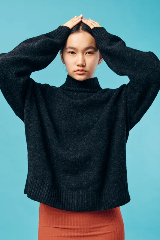 Young woman posing with hands on head wearing black sweater and orange skirt against blue background. Studio portrait of female model with serious expression and casual comfortable clothing.