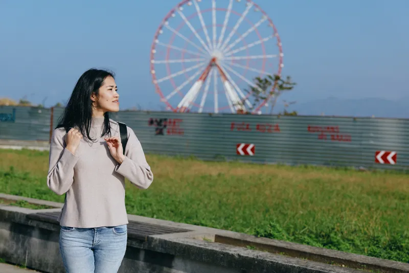 Woman with backpack strolling along a park path, with a large ferris wheel in the background, casual outfit under bright daylight, outdoor leisure and travel mood