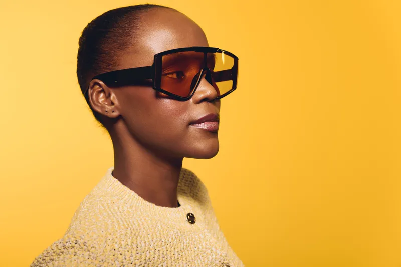 Profile of a confident African American woman with dark skin wearing oversized sunglasses and a textured sweater against a vibrant yellow background. Concept of fashion and style.