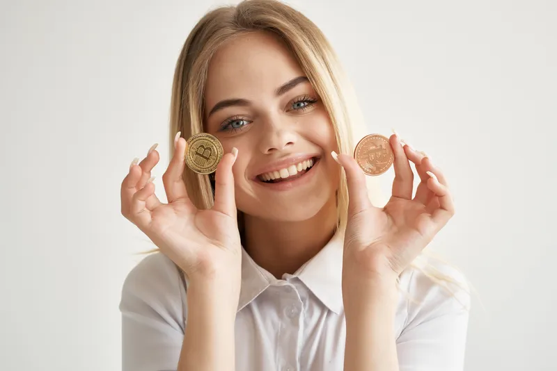 Жизнерадостная блондинка в белой рубашке держит криптовалюту Bitcoin возле своих глаз