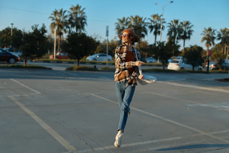 Woman jumping in a parking lot at golden hour, candid lifestyle portrait with sunglasses and casual jeans, joyful urban moment conveying authenticity, mindful living and emotional storytelling.