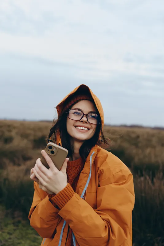 Woman lifestyle autumn outdoors portrait, smiling in orange hood jacket, holding smartphone for selfie, countryside field background, natural daylight, casual travel mood, open sky
