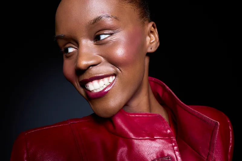 Smiling dark skinned African American woman in red leather jacket looks sideways with bright makeup and natural skin glow. Studio portrait with black background, concept of beauty and confidence.
