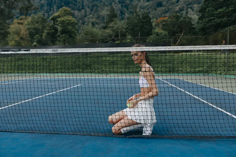 Young female tennis player in a white outfit kneeling on a blue tennis court, looking determined and focused, surrounded by nature and greenery