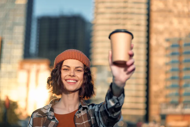 Woman holding coffee cup with smile in urban streets at golden hour, beanie and plaid jacket, candid lifestyle portrait with authenticity, mindful living and emotional storytelling.