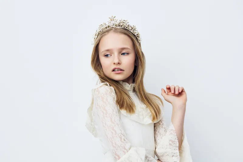 Young girl in a beautiful white dress with a crown, expressing elegance and grace against a soft pastel background Ideal for children s fashion and portrait photography