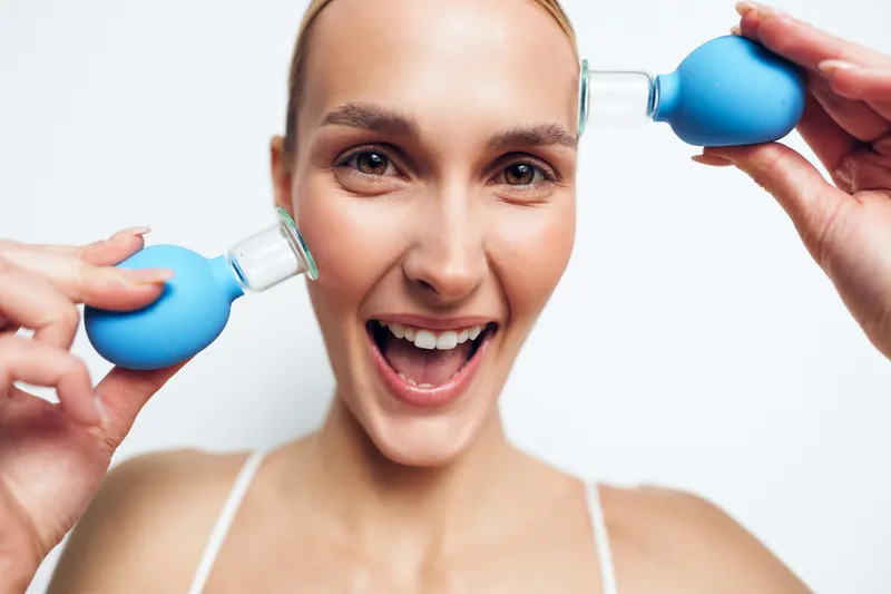 A cheerful woman demonstrates ear cleaning using two blue bulb syringes, showcasing personal hygiene and self care in a bright bathroom. Concept for wellness and grooming.