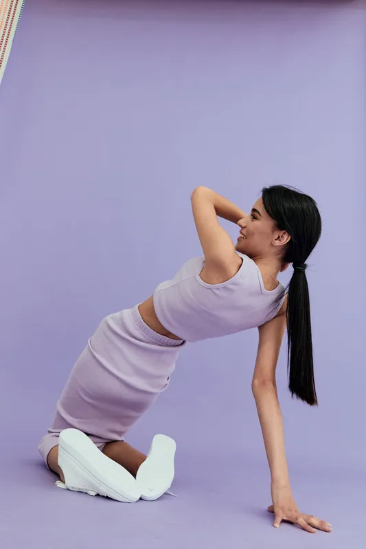 young woman in pastel outfit posing on purple background, wearing knitted crop top and skirt, casual and modern style with relaxed expression and long ponytail