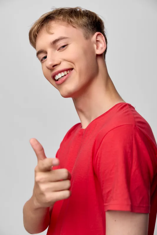 Smiling youth, pointing gesture, casual style, red shirt, confident pose, friendly vibe, studio shot, bright background, energetic and approachable scene highlighting modern lifestyle