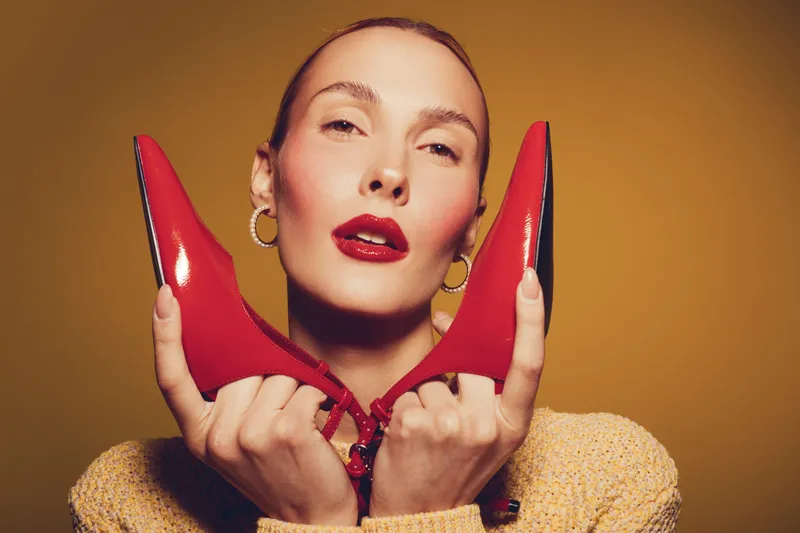 Stylish woman with red lipstick holds red high heel shoes near face on yellow background. Fashion concept with confident model, beauty, glamour, and modern style in studio lighting.
