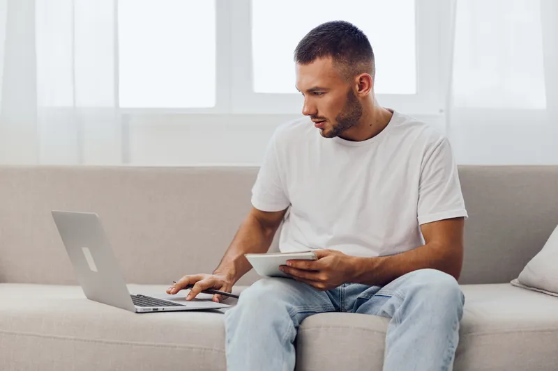 Focused man working on laptop while using tablet at home, showcasing a modern workspace and productivity in a bright environment.