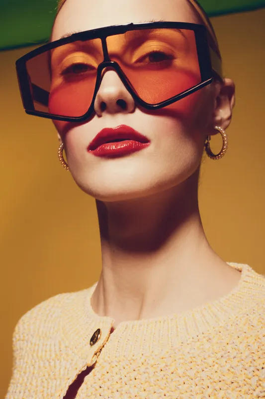 Stylish woman with oversized orange sunglasses and bold red lipstick poses confidently against yellow background. Fashion portrait with modern accessories and trendy makeup look.