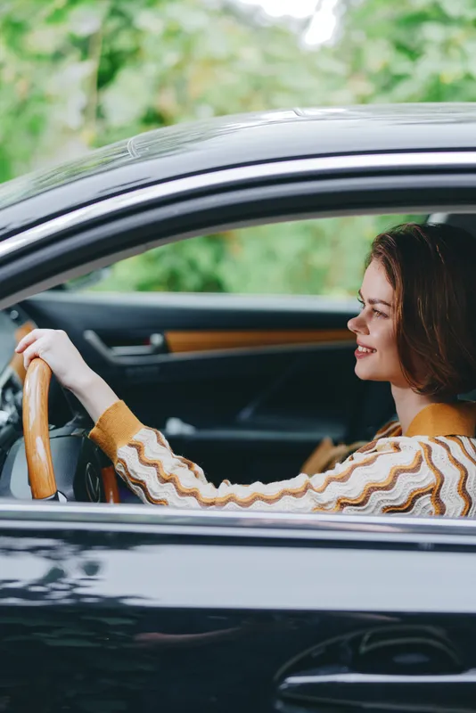 Smiling female driver sits behind the wheel of a sleek car, captured from the side with soft natural light and a blurred green background.