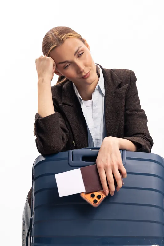 Tired woman in business attire leans on large suitcase holding phone and passport. Concept for travel fatigue, waiting, and business trip stress with white background.