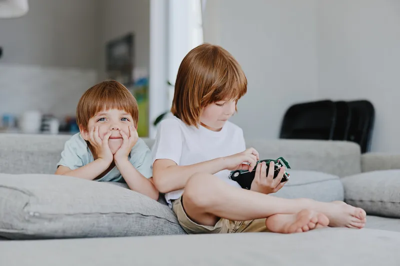 Two children playing at home, one boy resting his chin on his hands with a smile, while the other focuses on a toy. A cozy and inviting atmosphere surrounds them.