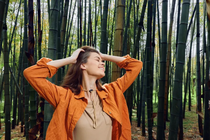 woman bamboo forest smile outdoor portrait nature relaxation and joy, female in orange jacket enjoying peaceful greenery, eyes closed and hands in hair during walk